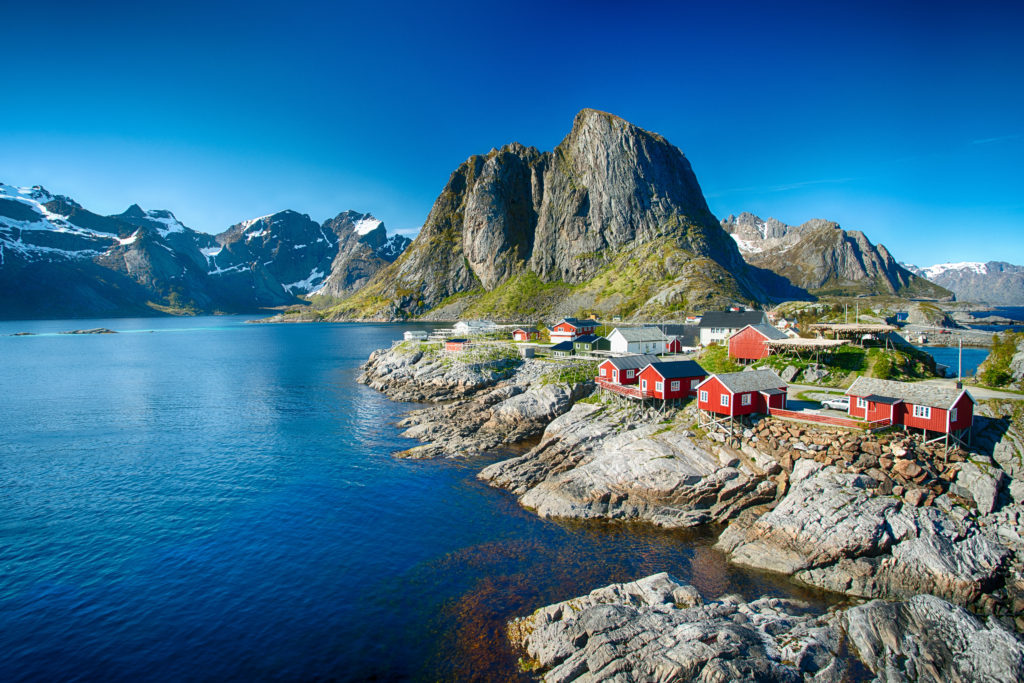 Ein kleines Dorf mit roten Fischerhütten auf den Lofoten, umgeben von blauen Fjorden und zum Teil schneebedeckten Bergen.
